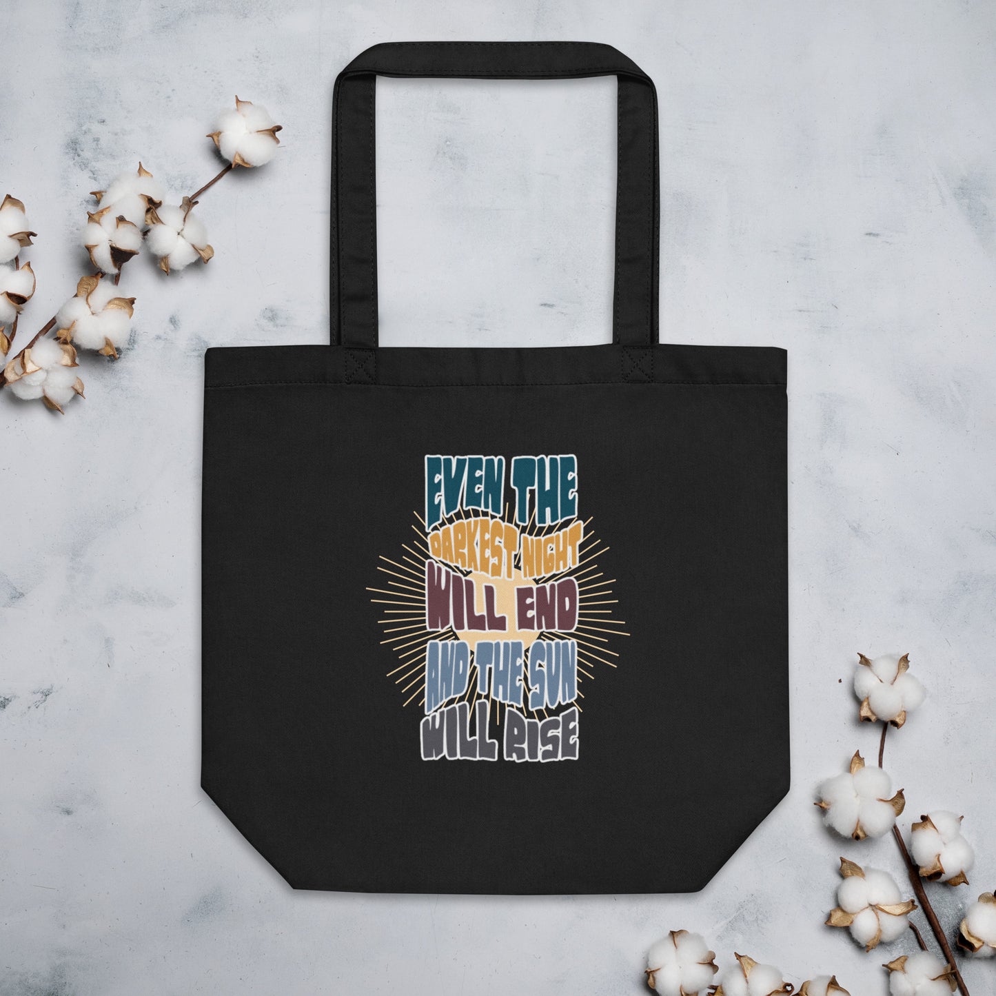 A black canvas tote bag featuring an inspirational quote in colorful, stylized typography with radiating yellow lines in the background. The text reads "EVEN THE DARKEST NIGHT WILL END AND THE SUN WILL RISE" with each phrase in a different color - blue, orange, purple, and light blue respectively. The bag has two black shoulder straps and a spacious rectangular shape.