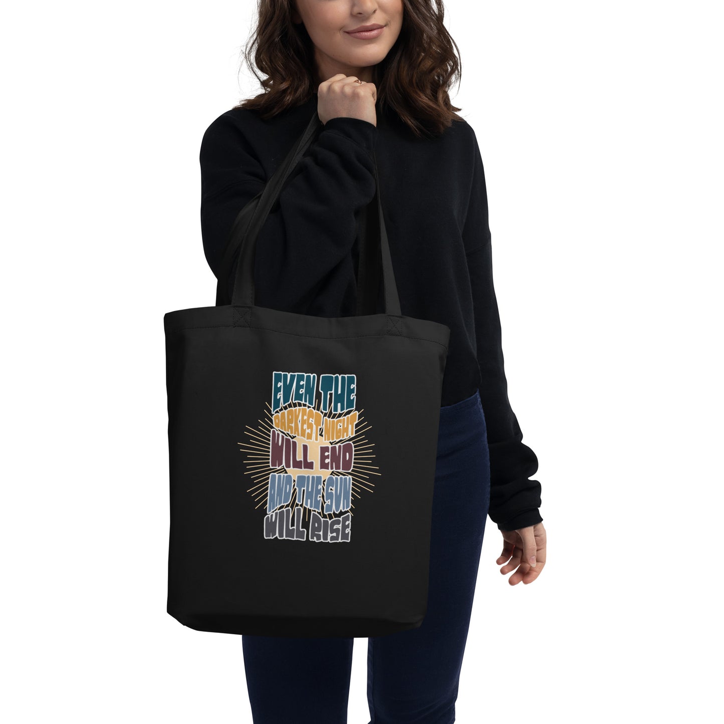 A woman holding up a black canvas tote bag featuring an inspirational quote in colorful, stylized typography with radiating yellow lines in the background. The text reads "EVEN THE DARKEST NIGHT WILL END AND THE SUN WILL RISE" with each phrase in a different color - blue, orange, purple, and light blue respectively. The bag has two black shoulder straps and a spacious rectangular shape.