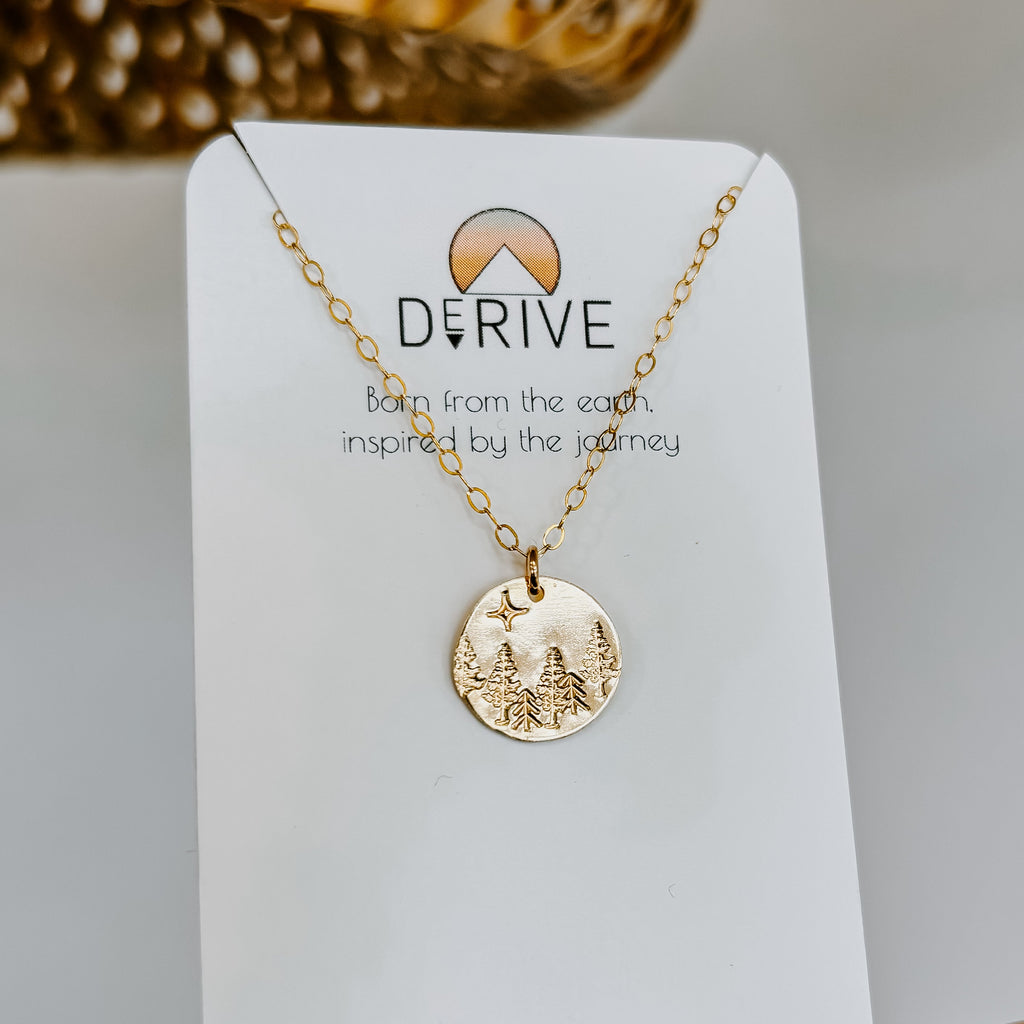 Gold mushroom pendant necklace on a delicate chain displayed on a white jewelry card with "DERIVE" branding and text "Born from the earth, inspired by the journey," photographed against a neutral background with dried flowers
