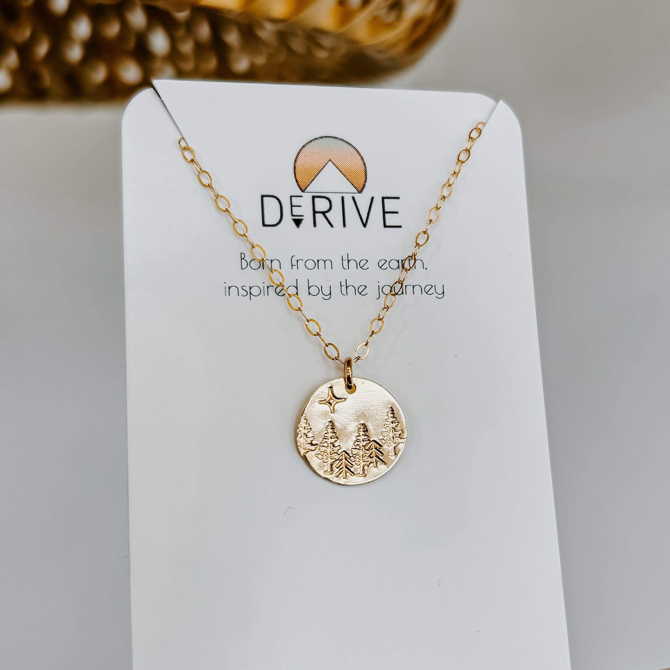 Gold mushroom pendant necklace on a delicate chain displayed on a white jewelry card with "DERIVE" branding and text "Born from the earth, inspired by the journey," photographed against a neutral background with dried flowers