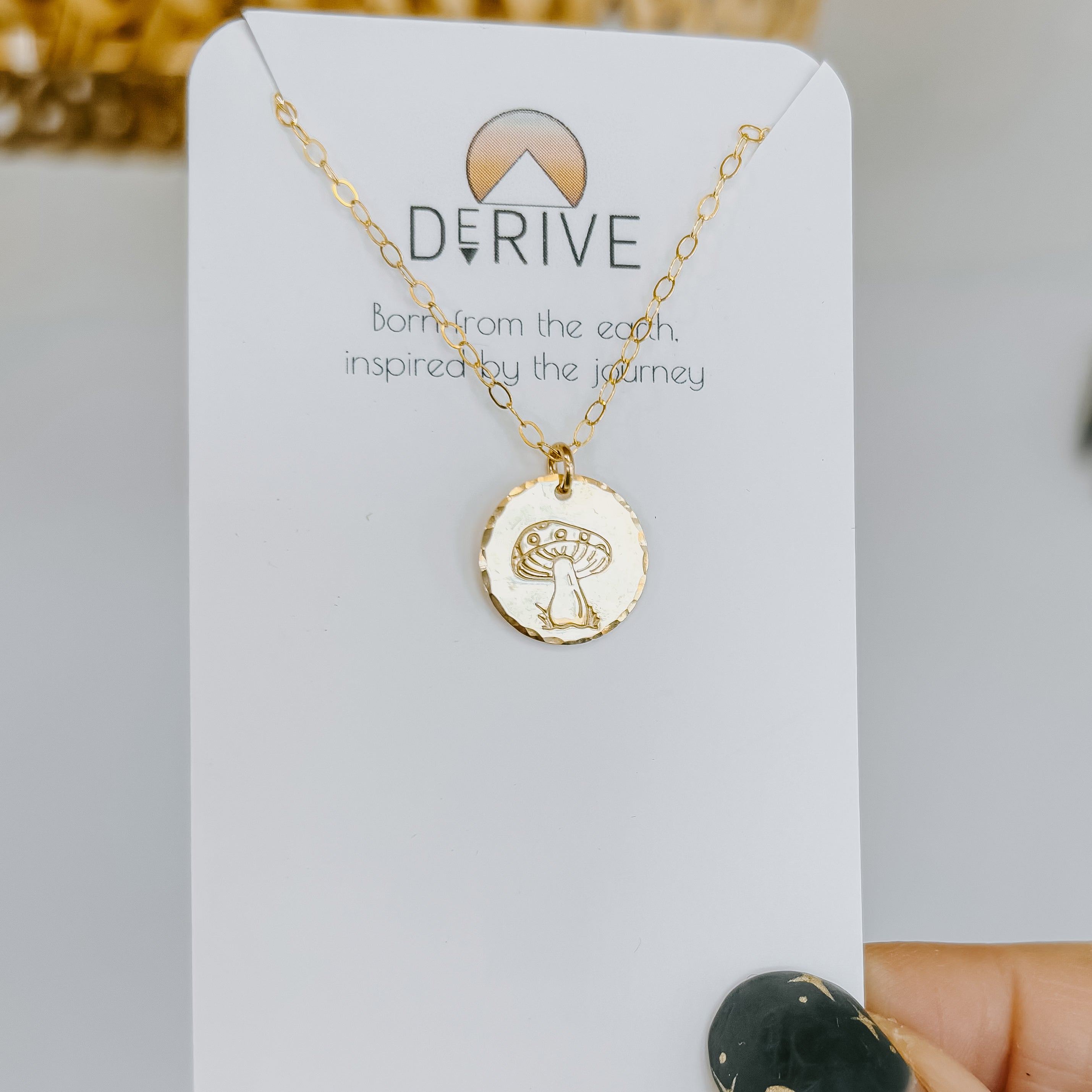 Gold mushroom pendant necklace on a delicate chain displayed on a white jewelry card with "DERIVE" branding and text "Born from the earth, inspired by the journey," photographed against a neutral background with dried flowers