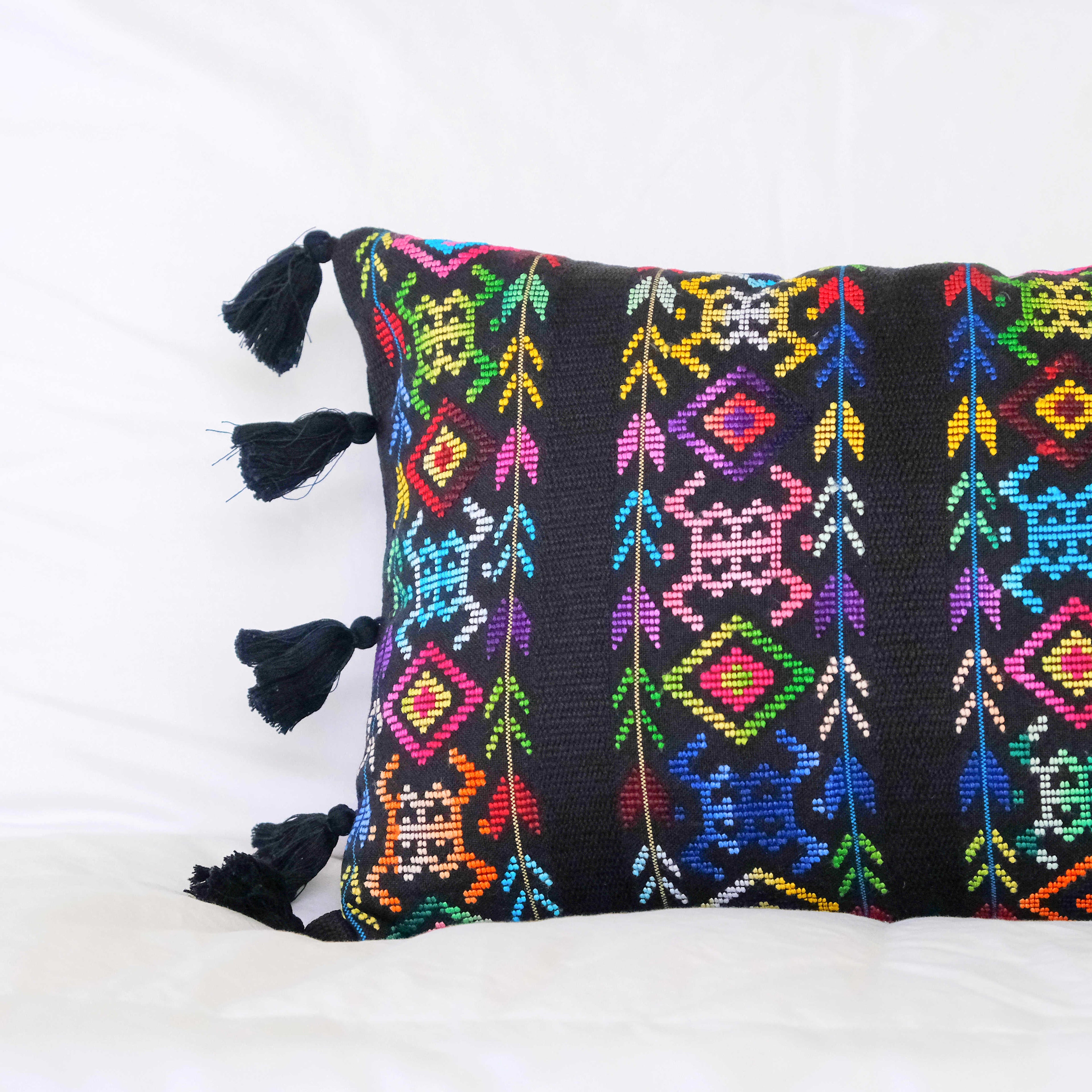 Hand woven lumbar pillow from Guatemala featuring intricate geometric patterns in vibrant colors including red, blue, pink, and white on a dark background, with bright blue tassel trim along both long edges, displayed against a white background