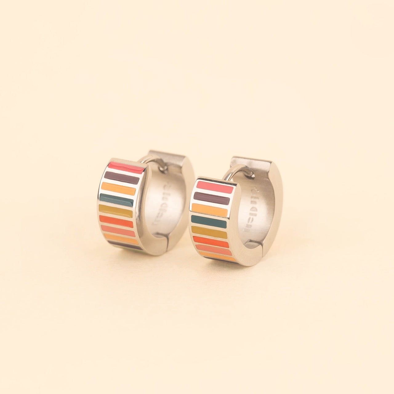 Pair of small gold-plated stainless steel hoop earrings with colorful horizontal stripes in pink, burgundy, orange, teal, and red enamel, photographed on a beige background