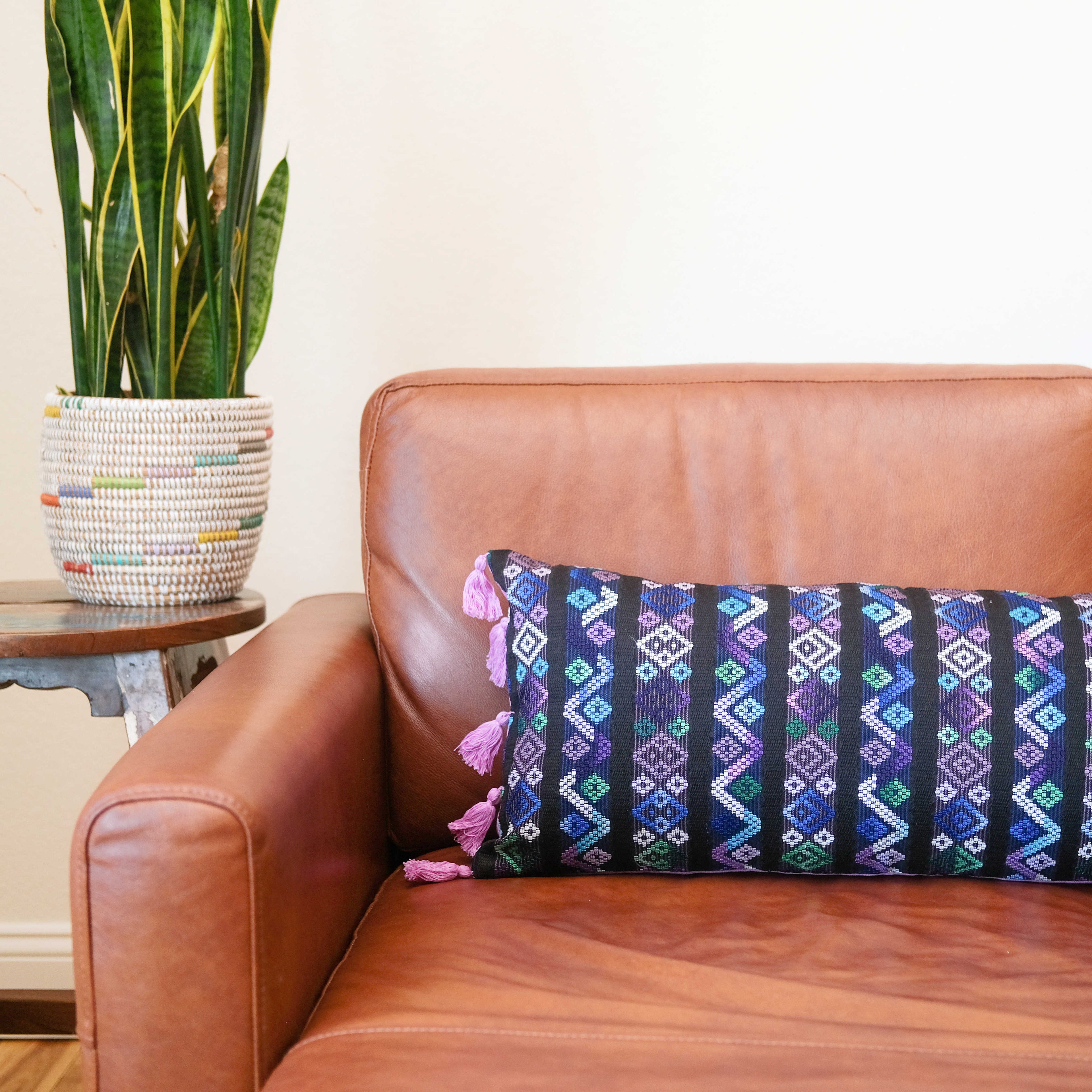 Hand woven lumbar pillow from Guatemala featuring intricate geometric patterns in vibrant colors including blue, pink, and white on a dark background, with pink tassel trim along both long edges, displayed against a white background
