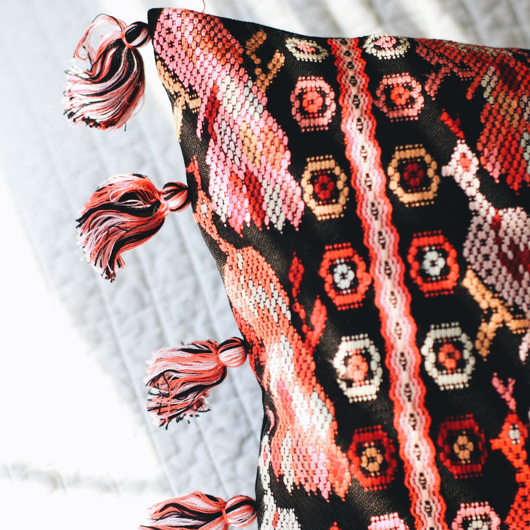 Hand woven lumbar pillow from Guatemala featuring intricate geometric patterns in vibrant colors including red, pink, and white on a dark background, with multicolor tassel trim along both long edges, displayed against a white background