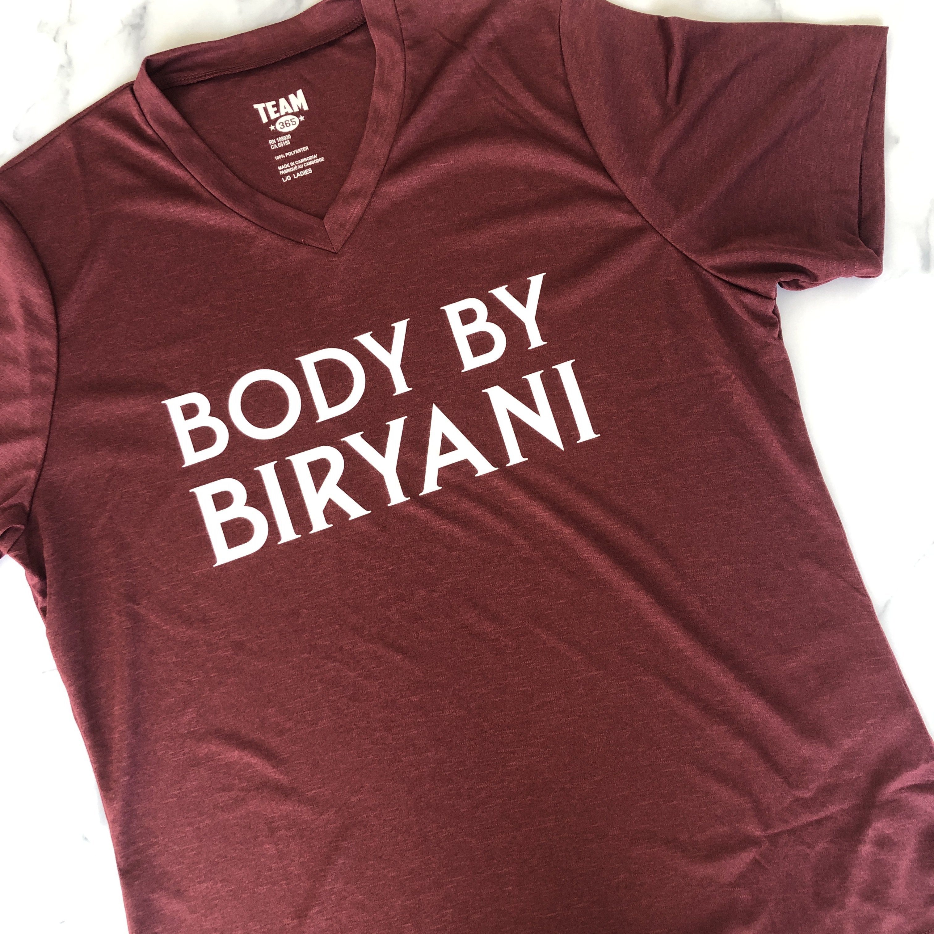 Burgundy maroon v-neck t-shirt featuring white text "BODY BY BIRYANI" in serif font, photographed on white marble surface