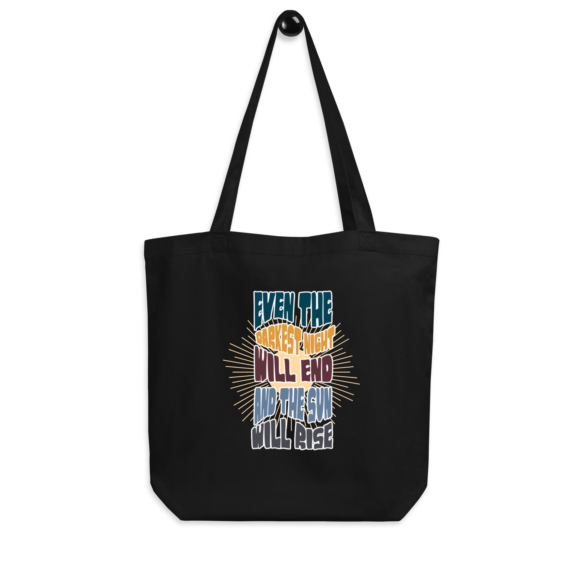 A black canvas tote bag featuring an inspirational quote in colorful, stylized typography with radiating yellow lines in the background. The text reads "EVEN THE DARKEST NIGHT WILL END AND THE SUN WILL RISE" with each phrase in a different color - blue, orange, purple, and light blue respectively. The bag has two black shoulder straps and a spacious rectangular shape.