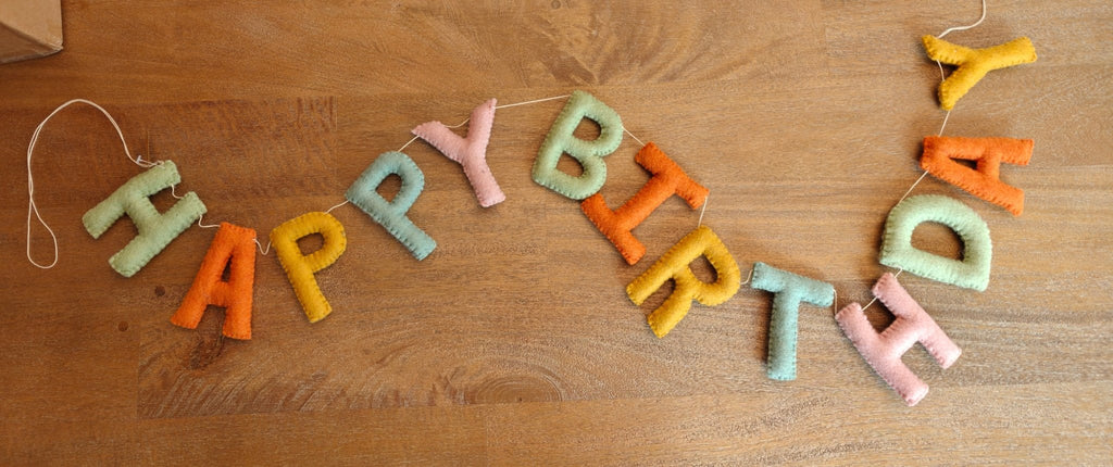A handmade felt 'HAPPY BIRTHDAY' banner garland in pastel colors, laid out on a wooden tabletop.
