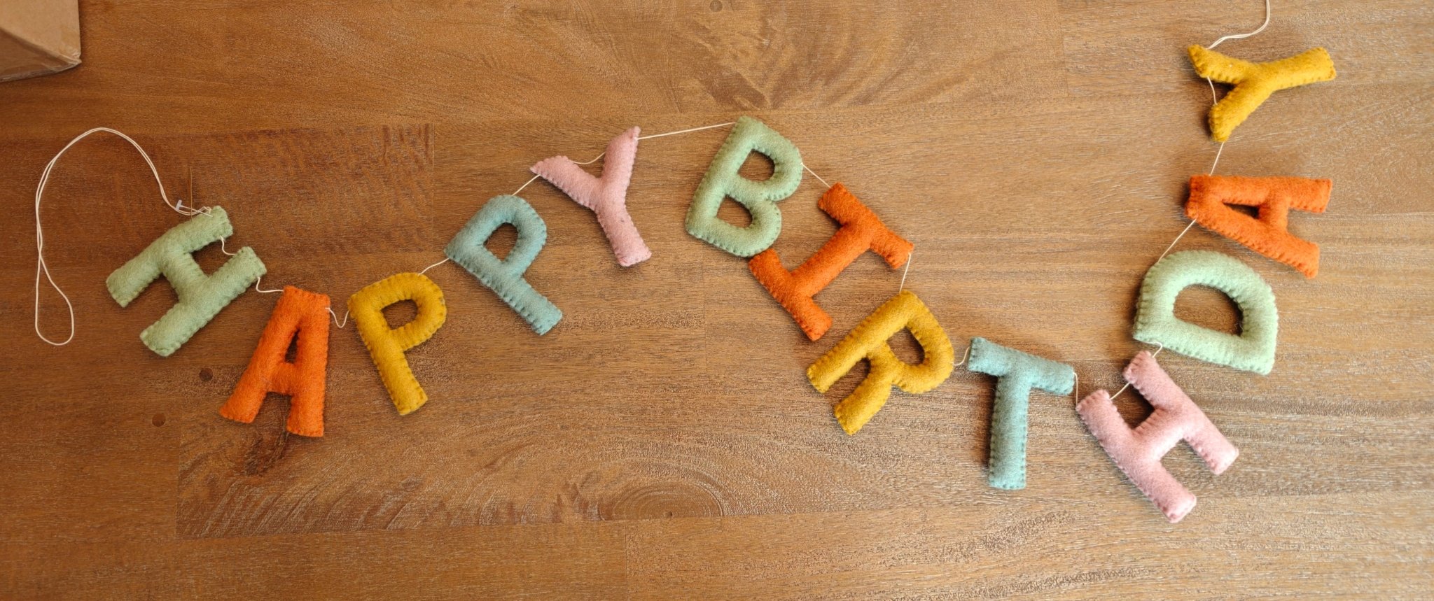 A handmade felt 'HAPPY BIRTHDAY' banner garland in pastel colors, laid out on a wooden tabletop.