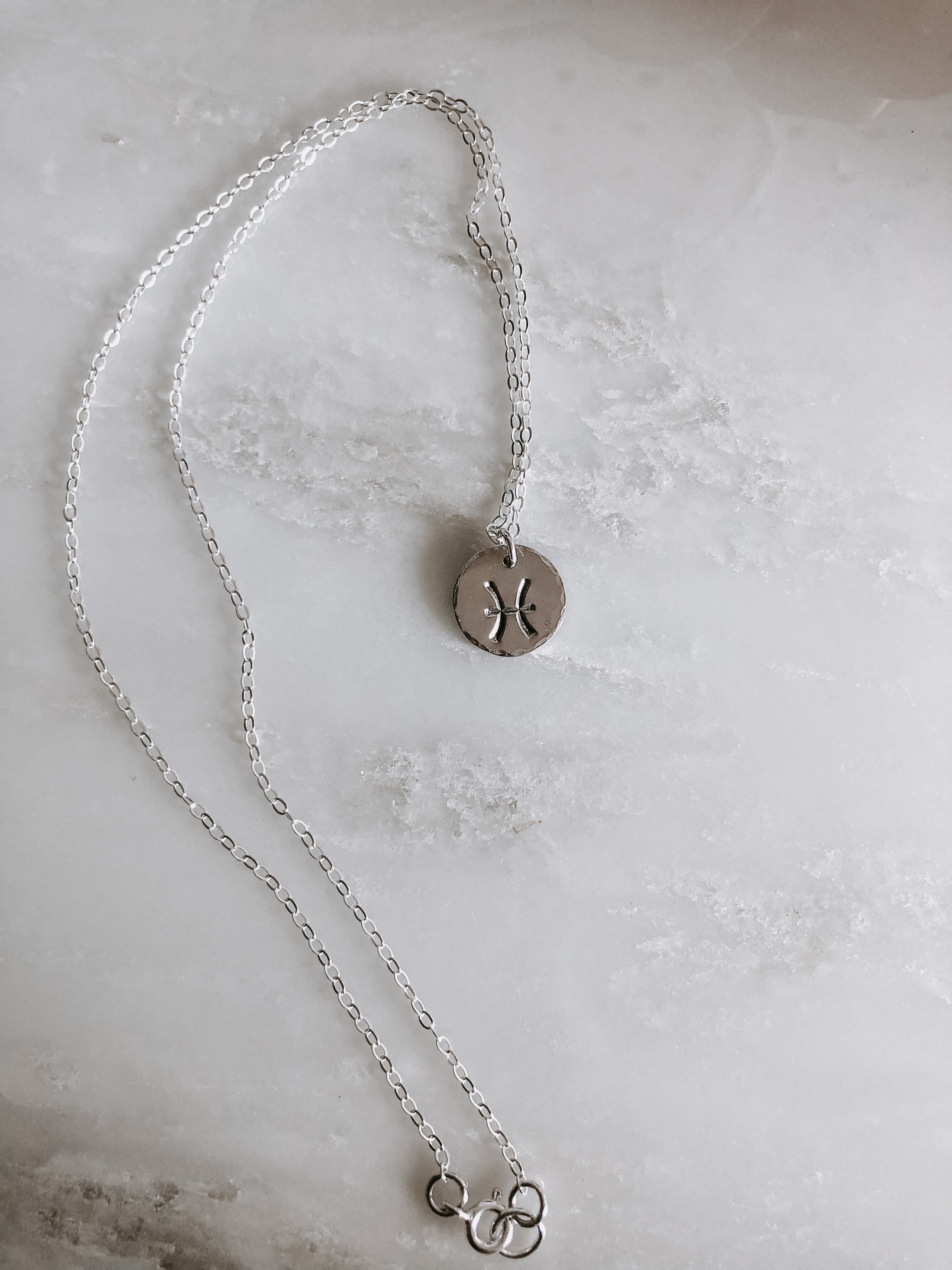 Collection of hand-stamped zodiac sign necklaces in gold and silver featuring circular pendants with astrological symbols including Aquarius, Scorpio, Taurus, Sagittarius, Cancer, Leo, Aries, Gemini, and Libra, displayed with delicate chains on a marble surface