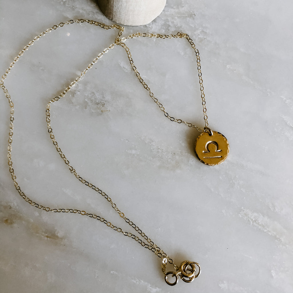 Collection of hand-stamped zodiac sign necklaces in gold and silver featuring circular pendants with astrological symbols including Aquarius, Scorpio, Taurus, Sagittarius, Cancer, Leo, Aries, Gemini, and Libra, displayed with delicate chains on a marble surface