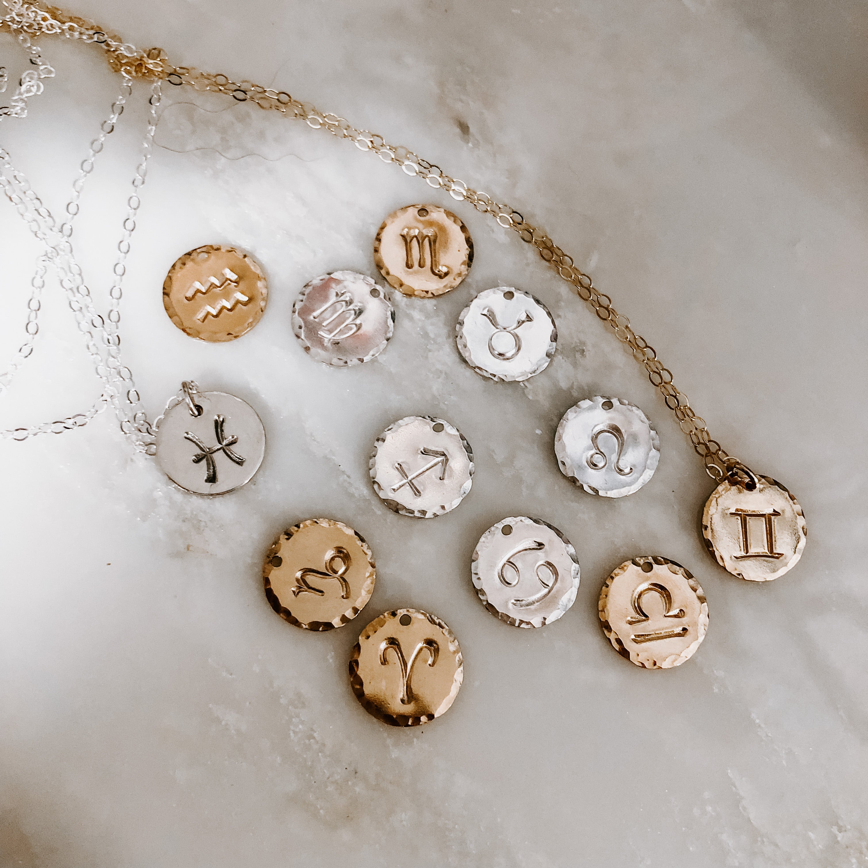 Collection of hand-stamped zodiac sign necklaces in gold and silver featuring circular pendants with astrological symbols including Aquarius, Scorpio, Taurus, Sagittarius, Cancer, Leo, Aries, Gemini, and Libra, displayed with delicate chains on a marble surface