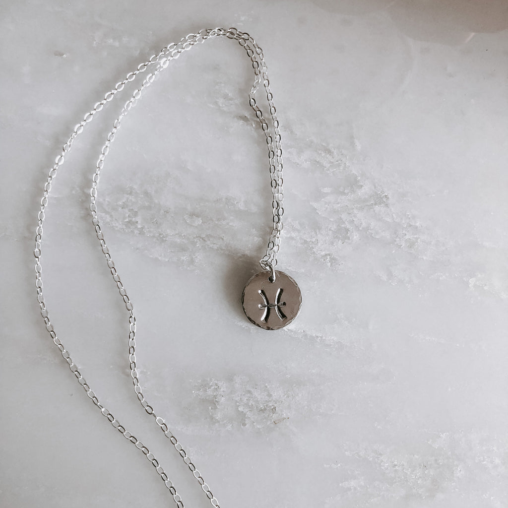 Collection of hand-stamped zodiac sign necklaces in gold and silver featuring circular pendants with astrological symbols including Aquarius, Scorpio, Taurus, Sagittarius, Cancer, Leo, Aries, Gemini, and Libra, displayed with delicate chains on a marble surface