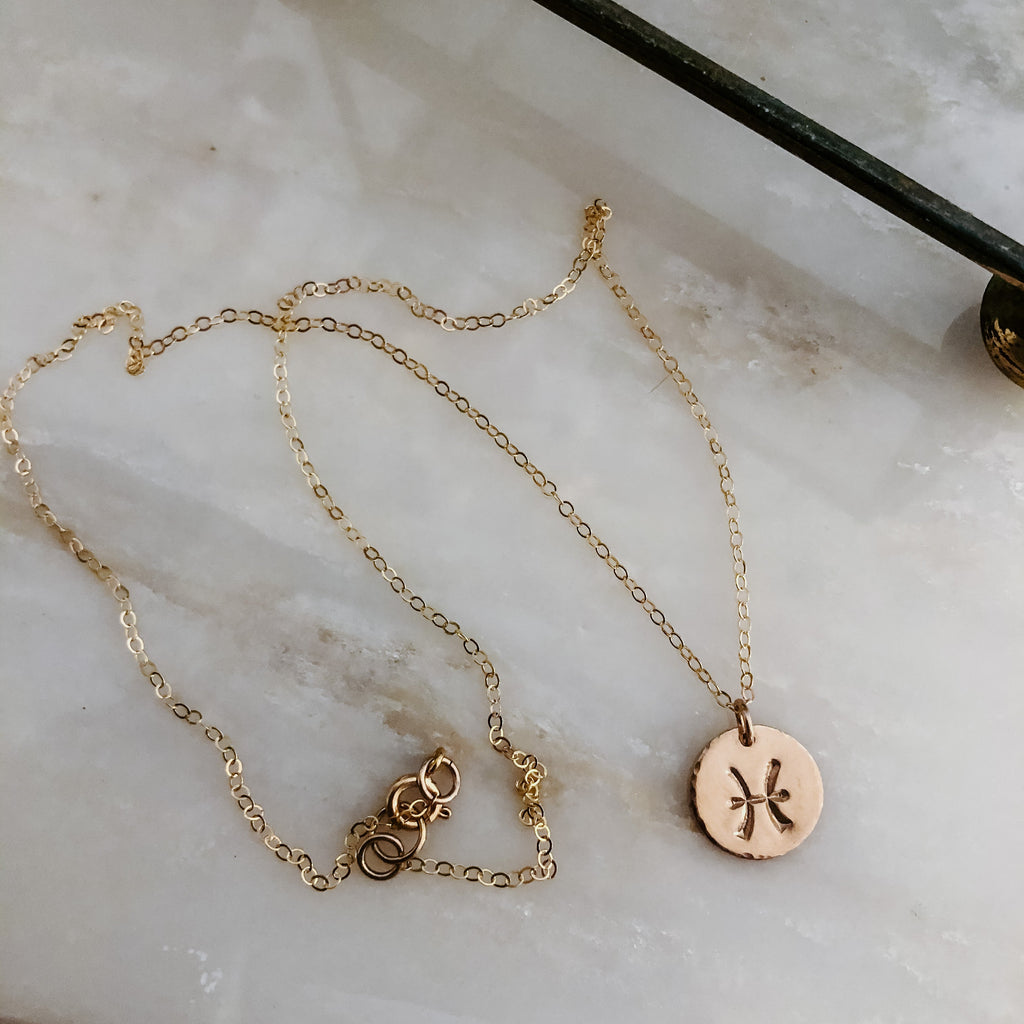 Collection of hand-stamped zodiac sign necklaces in gold and silver featuring circular pendants with astrological symbols including Aquarius, Scorpio, Taurus, Sagittarius, Cancer, Leo, Aries, Gemini, and Libra, displayed with delicate chains on a marble surface