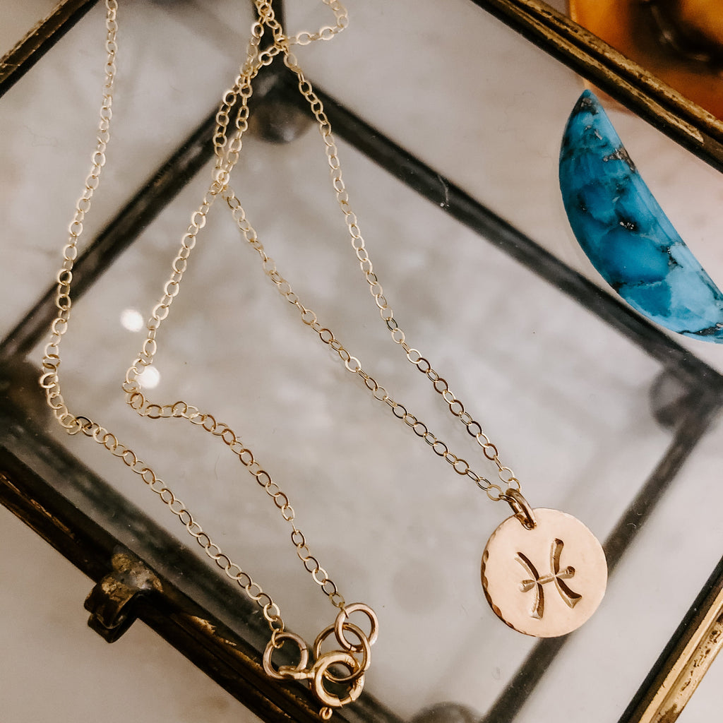 Collection of hand-stamped zodiac sign necklaces in gold and silver featuring circular pendants with astrological symbols including Aquarius, Scorpio, Taurus, Sagittarius, Cancer, Leo, Aries, Gemini, and Libra, displayed with delicate chains on a marble surface