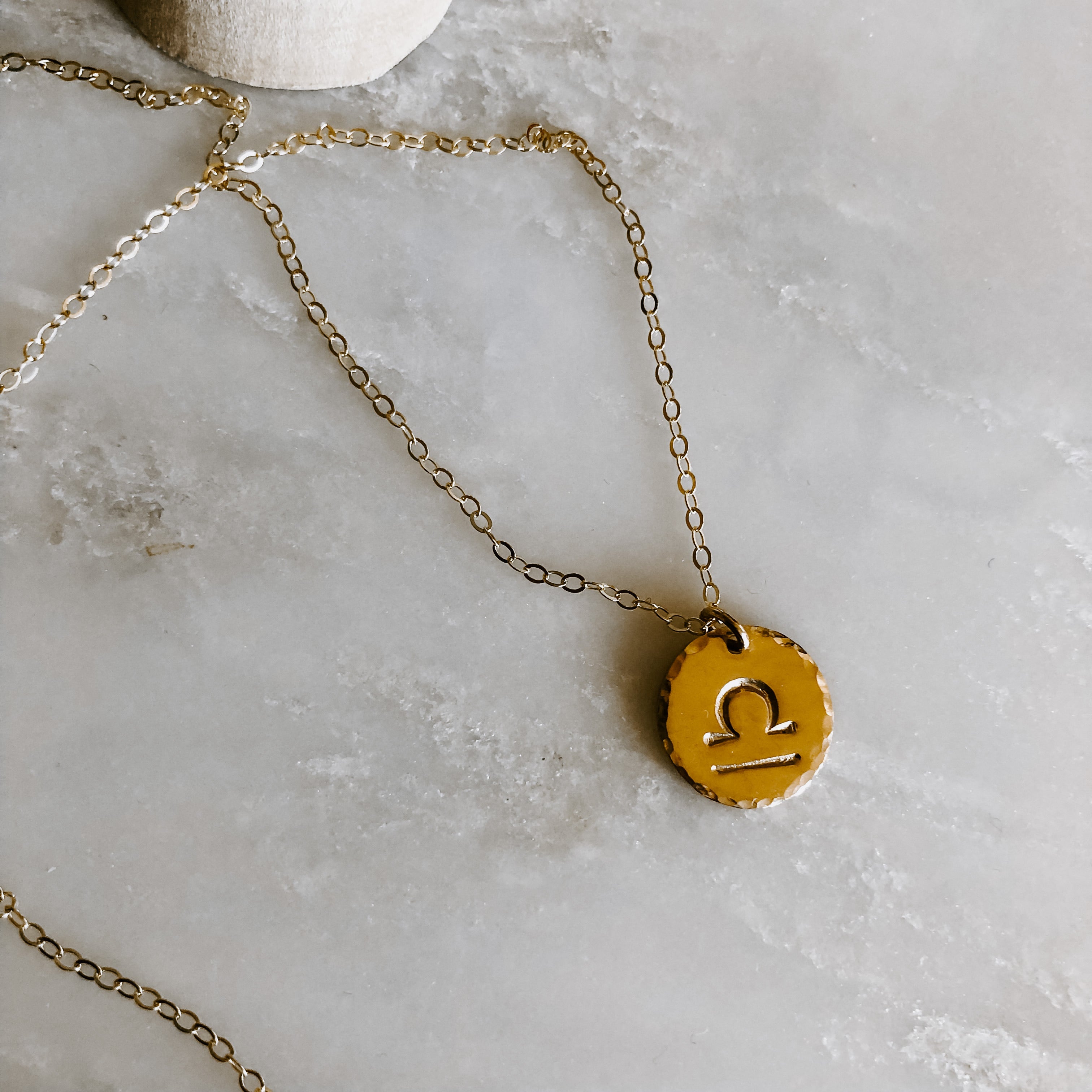 Collection of hand-stamped zodiac sign necklaces in gold and silver featuring circular pendants with astrological symbols including Aquarius, Scorpio, Taurus, Sagittarius, Cancer, Leo, Aries, Gemini, and Libra, displayed with delicate chains on a marble surface