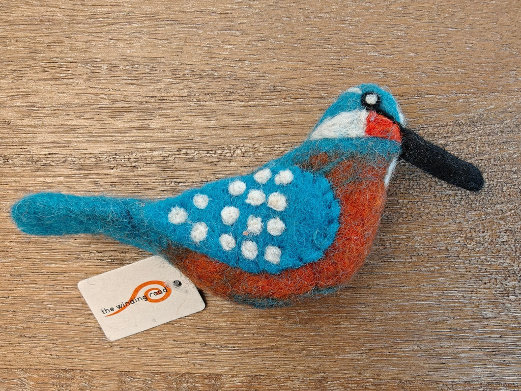 A handcrafted felted wool bird ornament, a kingfisher, with a blue back, white spots, orange-red belly, and black beak, resting on a wooden surface with a small paper tag attached.