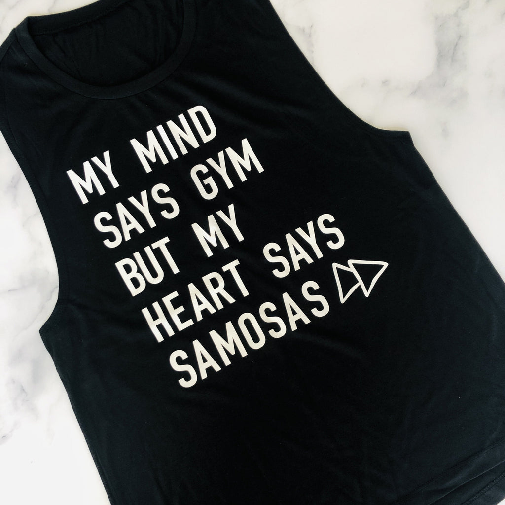 Black sleeveless tank top featuring white text "MY MIND SAYS GYM BUT MY HEART SAYS SAMOSAS" with arrow icon, photographed on white marble surface