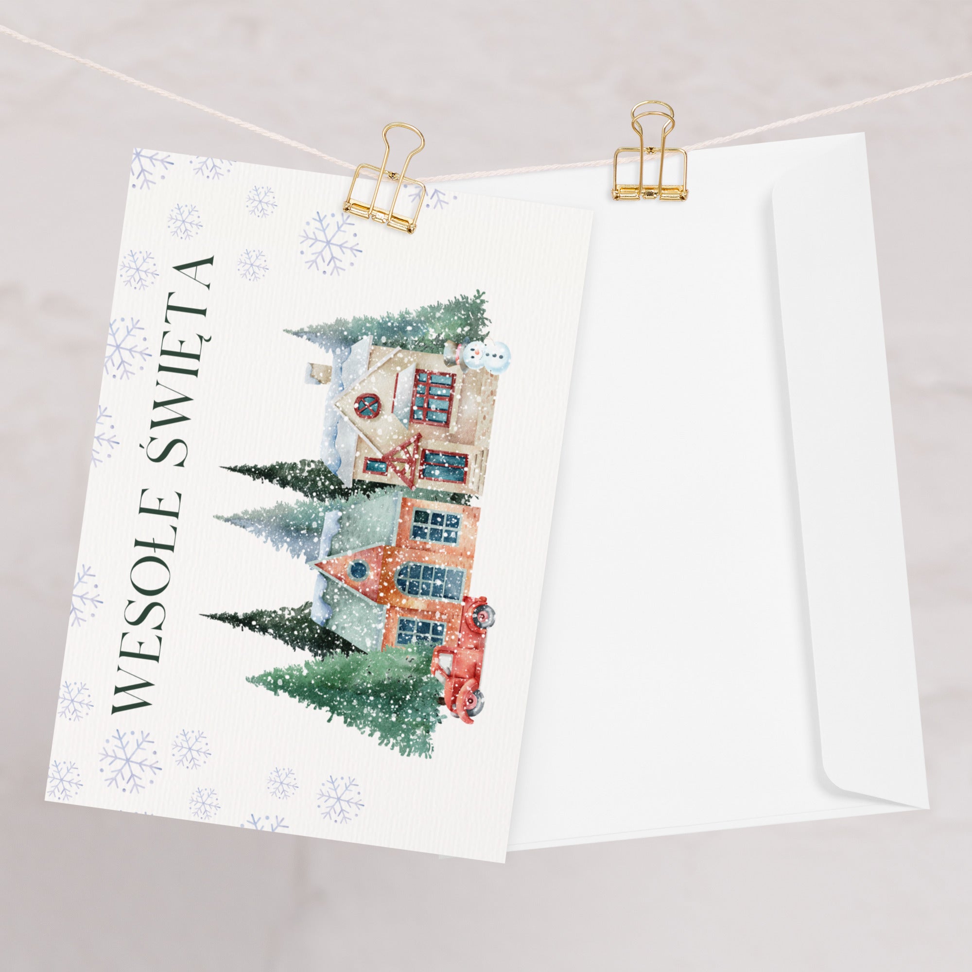 Polish Christmas greeting card featuring "Wesołe Święta" text with watercolor illustration of snowy village scene with cottages, pine trees, snowman, and vintage truck surrounded by delicate snowflake pattern, displayed with white envelope
