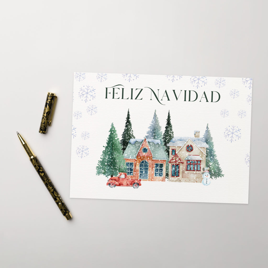 Spanish Christmas greeting card featuring "Feliz Navidad" text with watercolor illustration of snowy village scene with cottages, pine trees, snowman, and vintage truck surrounded by delicate snowflake pattern, displayed with white envelope