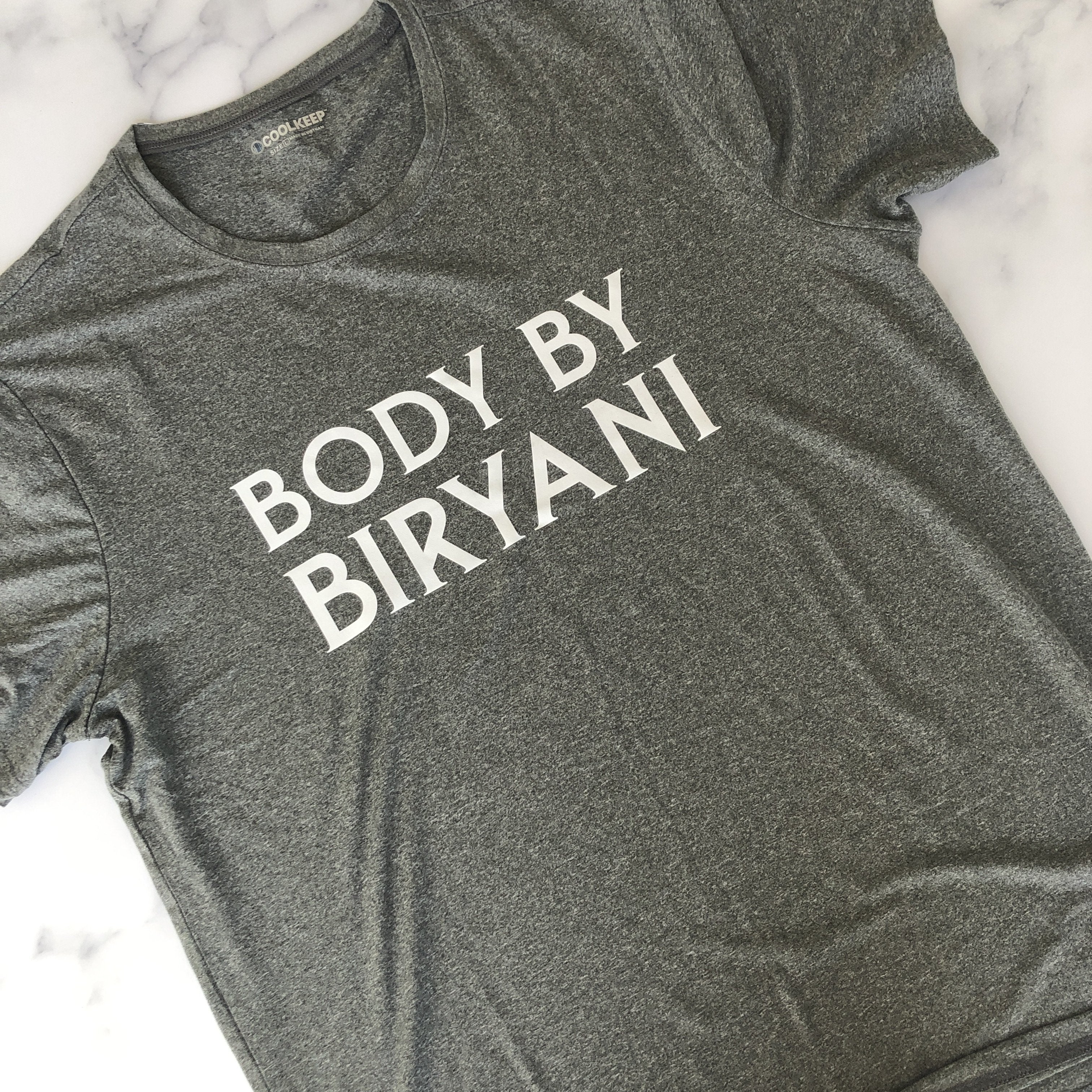 Dark heather gray t-shirt featuring white text "BODY BY BIRYANI" in bold uppercase serif font, photographed on white marble surface